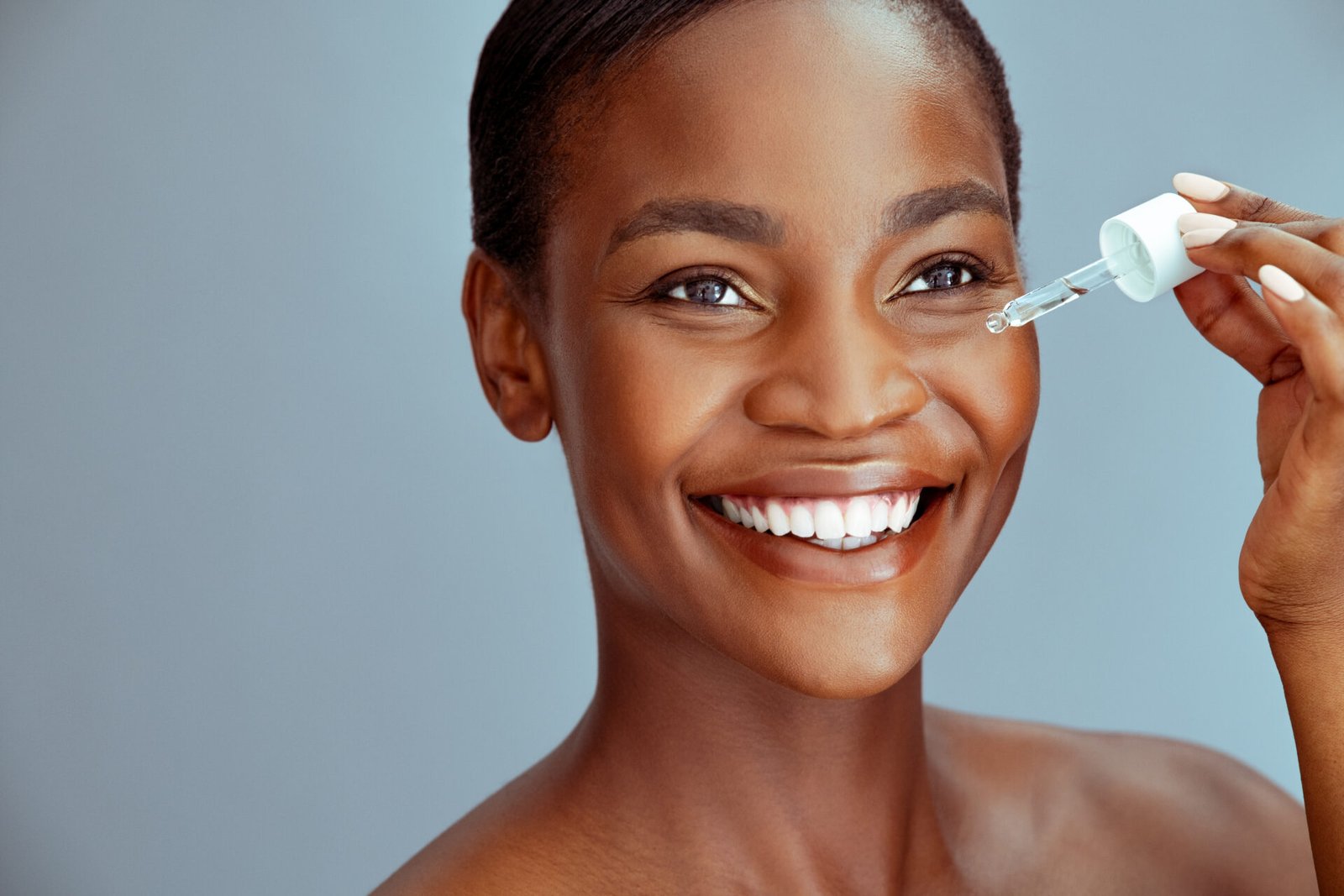 Portrait of african american woman applying liquid cosmetic oil for facial treatment against blue background with copy space. Beauty black middle aged woman with natural makeup holding glass dropper. Close up face of mature lady applying serum around dark circle eyes to combat wrinkles.