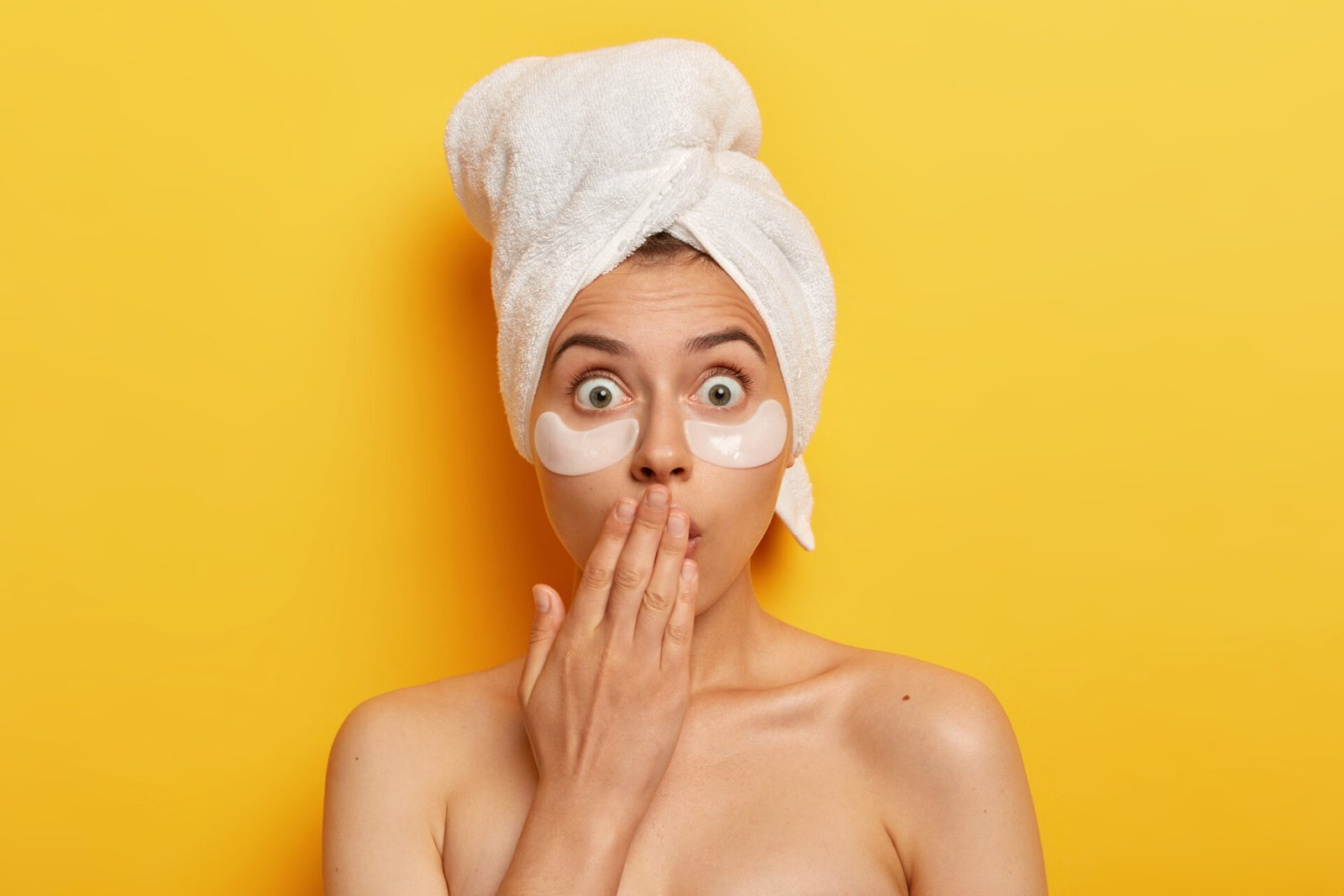 Shocked spa woman popping eyes at camera, horrified by terrible relevation, nourishes undereye skin with beauty patches, wears wrapped towel on head, has anti wrinkle procedure, being naked.