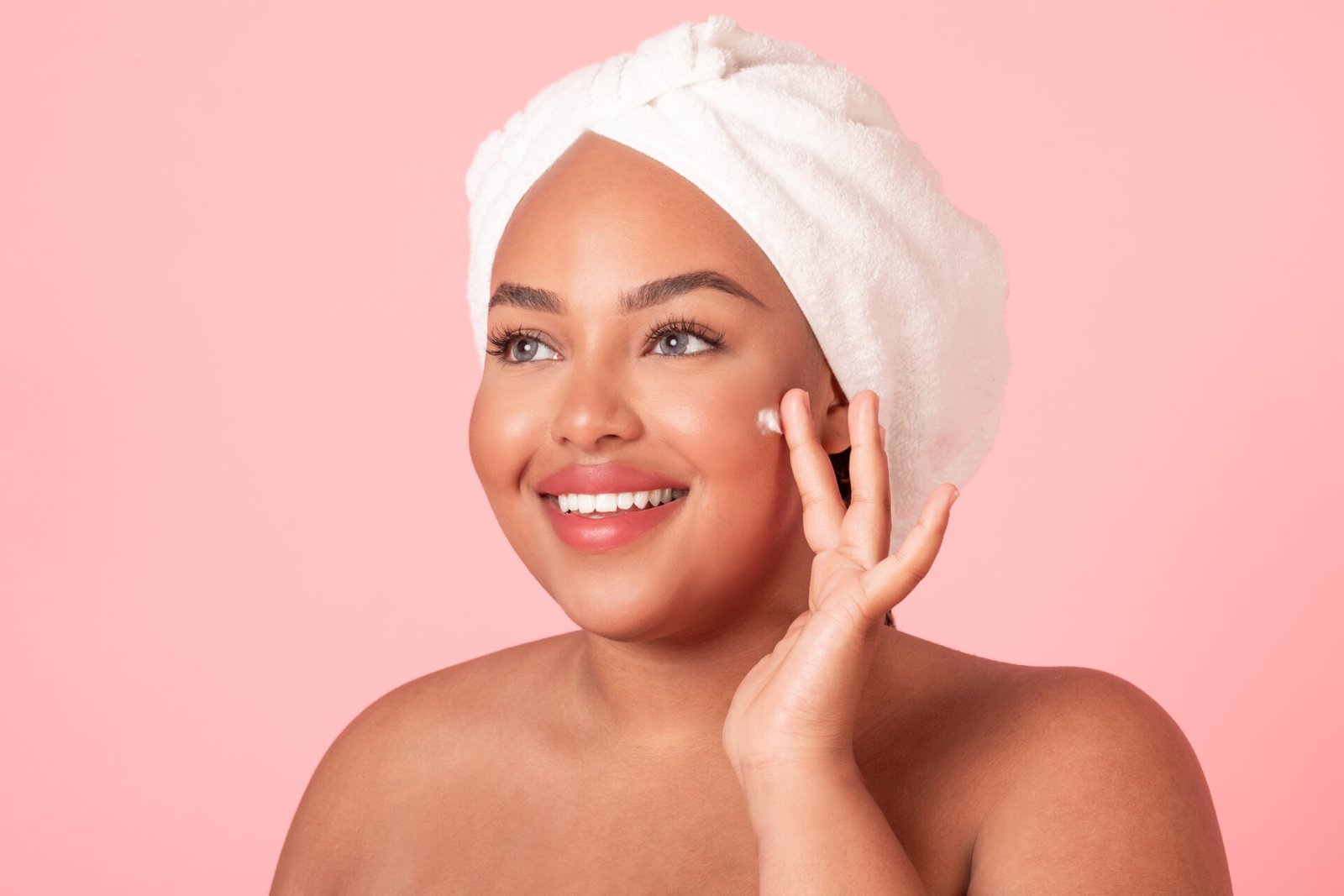 Skin care concept. Closeup portrait of black plus size lady applying moisturizing cream on face, posing over pink studio background and smiling
