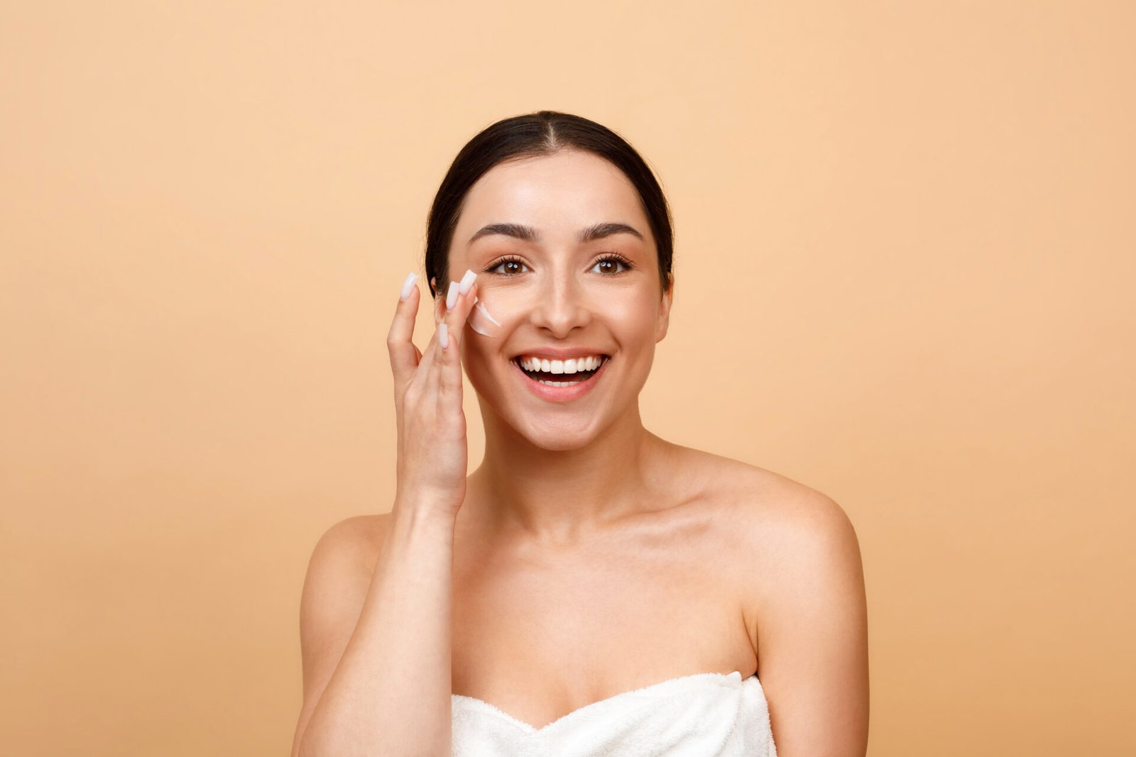 Skincare Routine. Happy Young Indian Woman Applying Moisturizing Cream On Face, Smiling Beautiful Hindu Female Wrapped In Towel Rubbing Nourishing Lotion While Posing On Beige Background, Copy Space